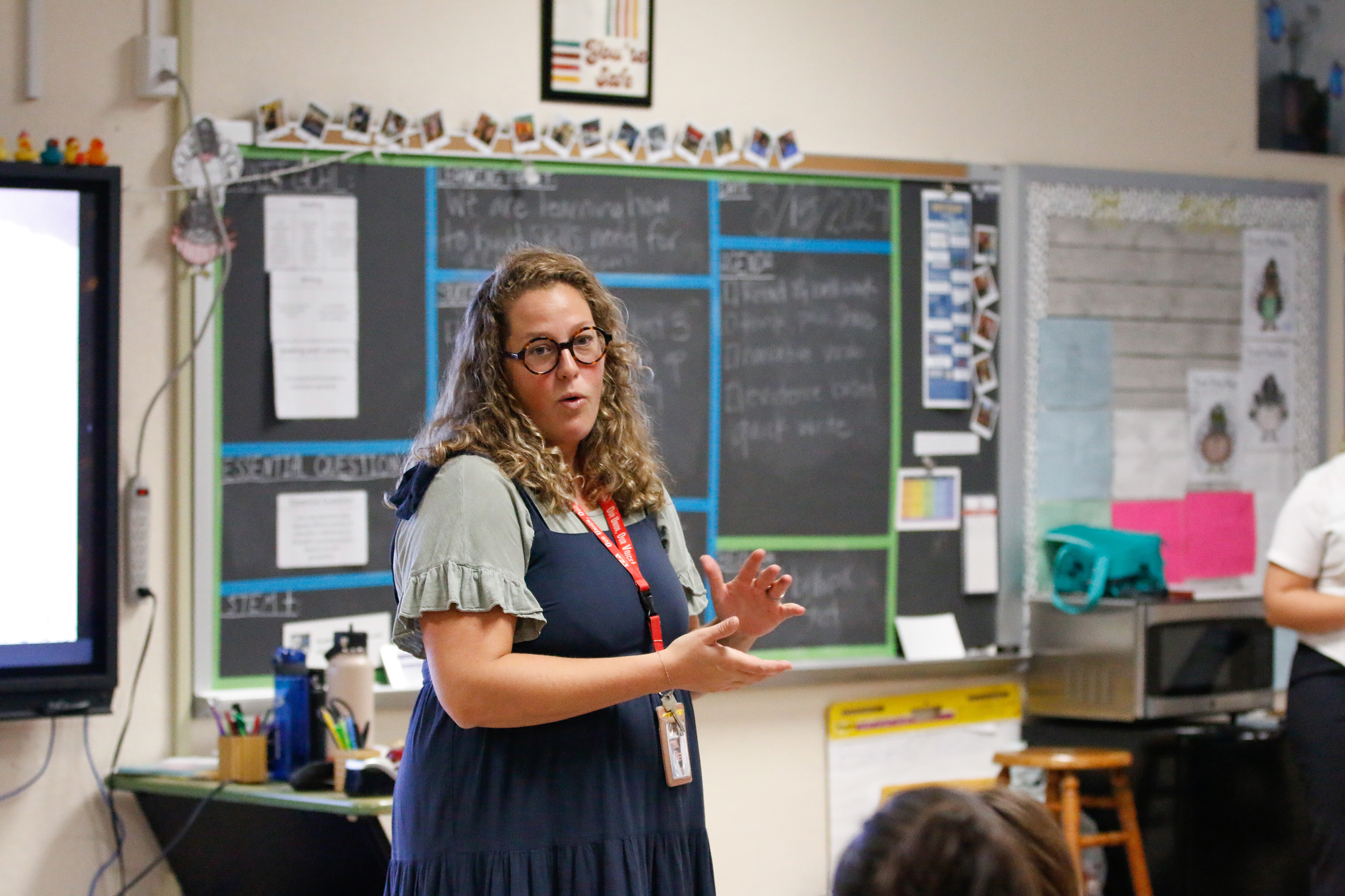 A teacher with glasses and curly blonde hair stands at the front of her classroom talking to families