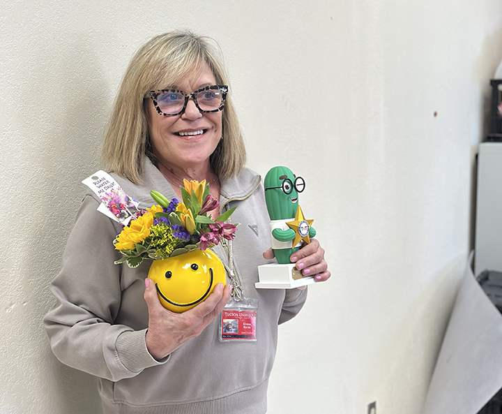 A woman with short blonde hair and glasses smiles holding up a yellow smiley planter and saguaro plushie