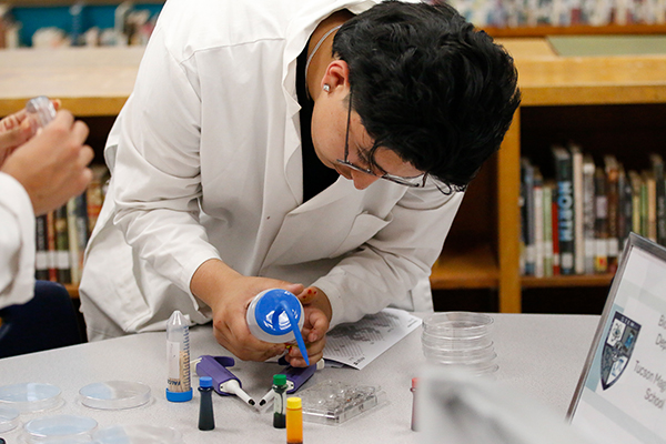 A teen boy in a white lab coat pours colored liquid into a tray