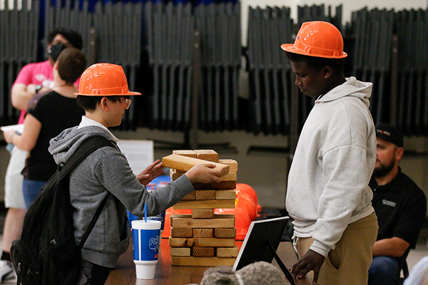 A boy in an orange hard hat lays giant Jenga pieces on top of each other as a teen boy in an orange hard hat watches behind the table
